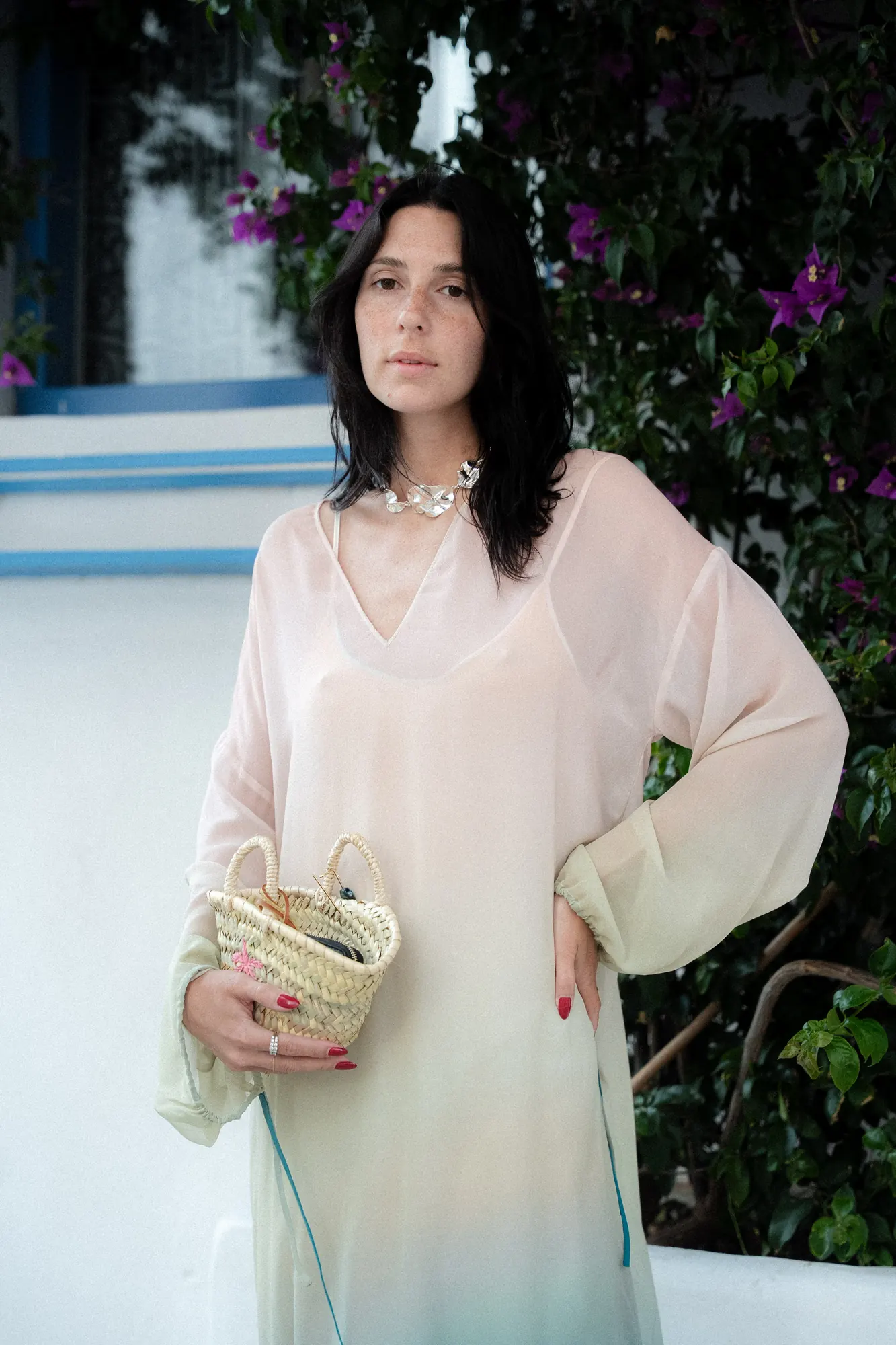 Ospite in abito di seta sfumato rosa e verde con borsa in paglia, davanti a un muro bianco calce e a una bouganville fiorita / Guest in a pink and green ombré silk dress holding a straw bag, in front of a whitewashed wall and a flowering bougainvillea