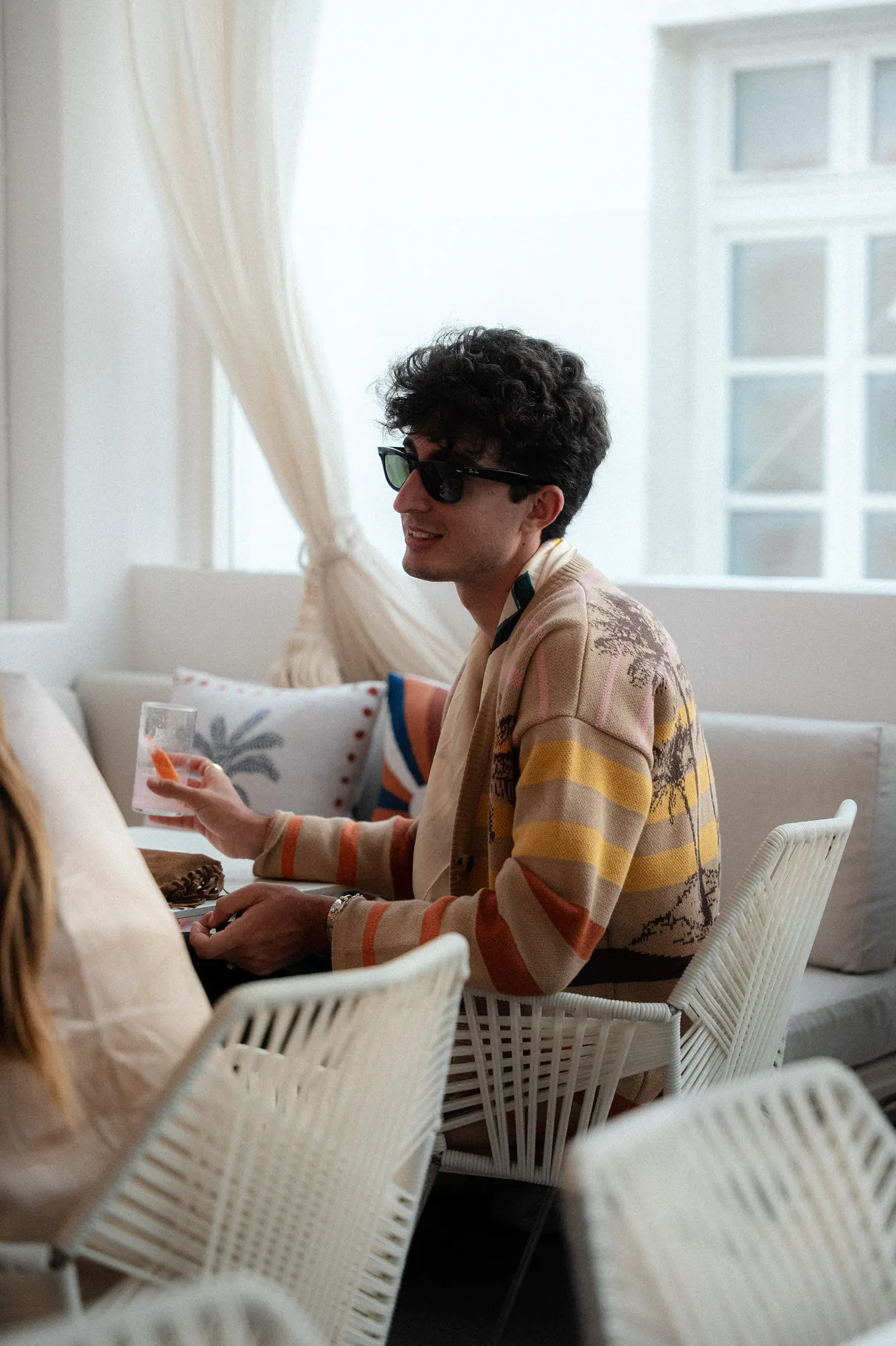 Ospite con occhiali da sole e cardigan in maglia a righe e palme seduto in una lounge bianca con un drink in mano / Guest in sunglasses and a striped palm-motif knit cardigan seated in a white lounge holding a drink