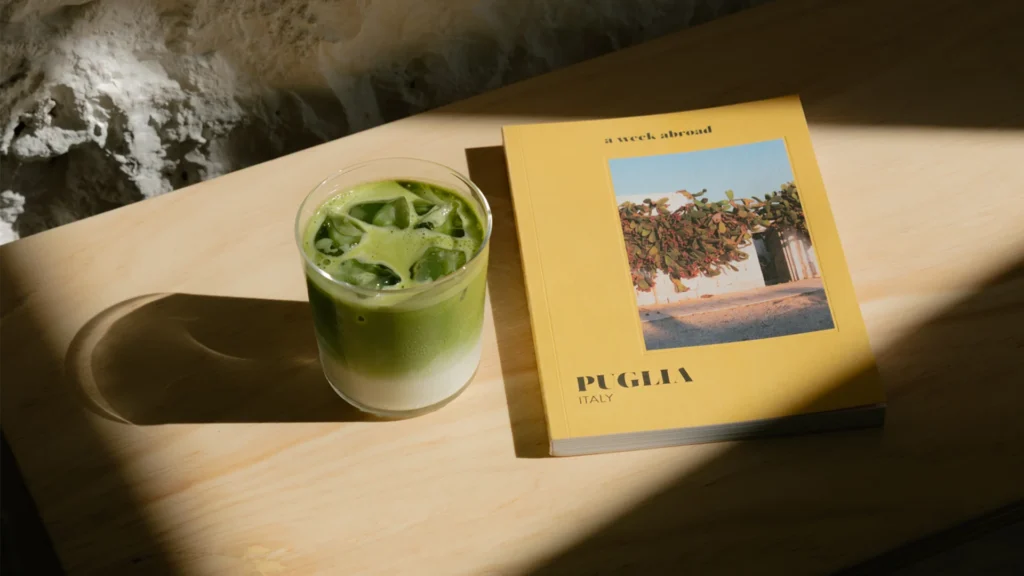 Bicchiere di matcha latte freddo accanto a una guida di viaggio gialla "A Week Abroad – Puglia, Italy" su un tavolo di legno chiaro illuminato dal sole / Glass of iced matcha latte next to a yellow travel guide "A Week Abroad – Puglia, Italy" on a sunlit light wood table