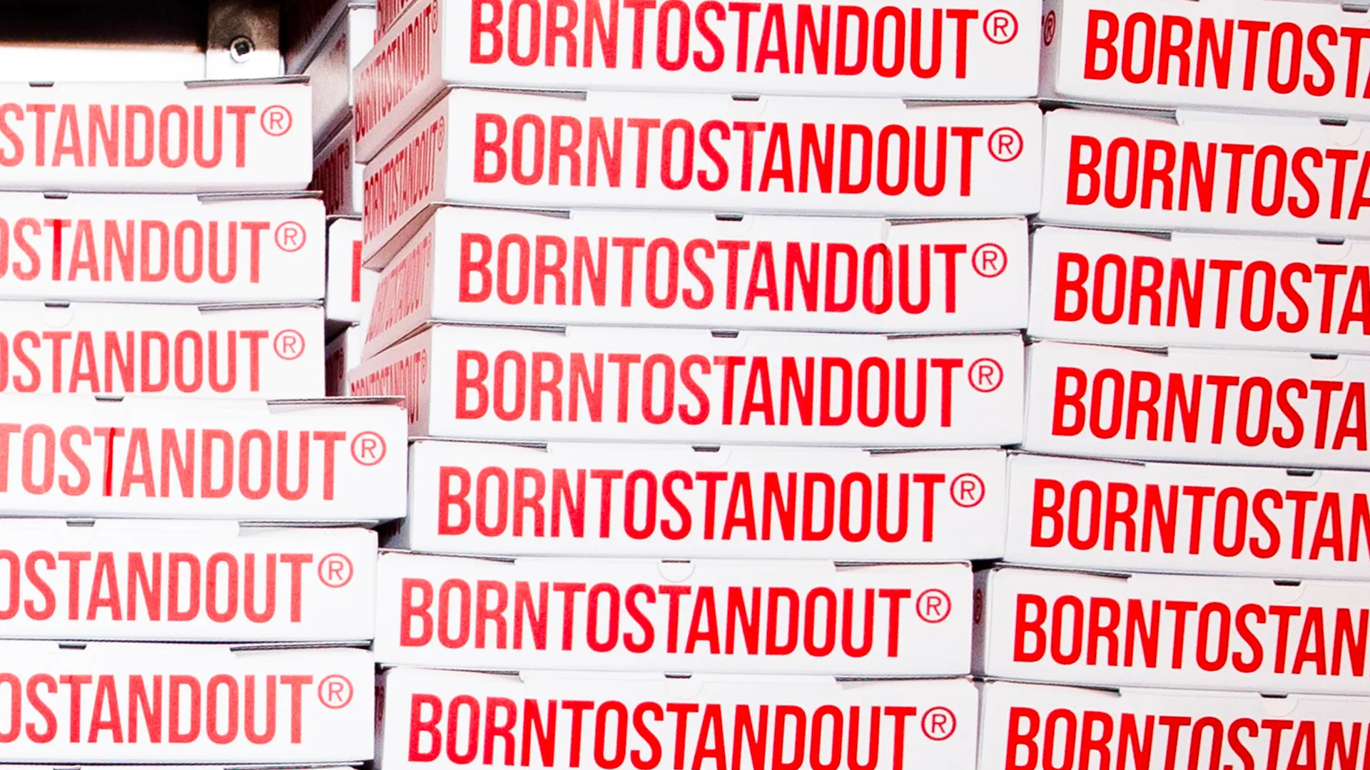 Cataste di scatole per pizza bianche con la scritta rossa BORNTOSTANDOUT ripetuta, allestimento del Craving Club / Stacks of white pizza boxes printed with the red BORNTOSTANDOUT logo repeated, Craving Club event setup