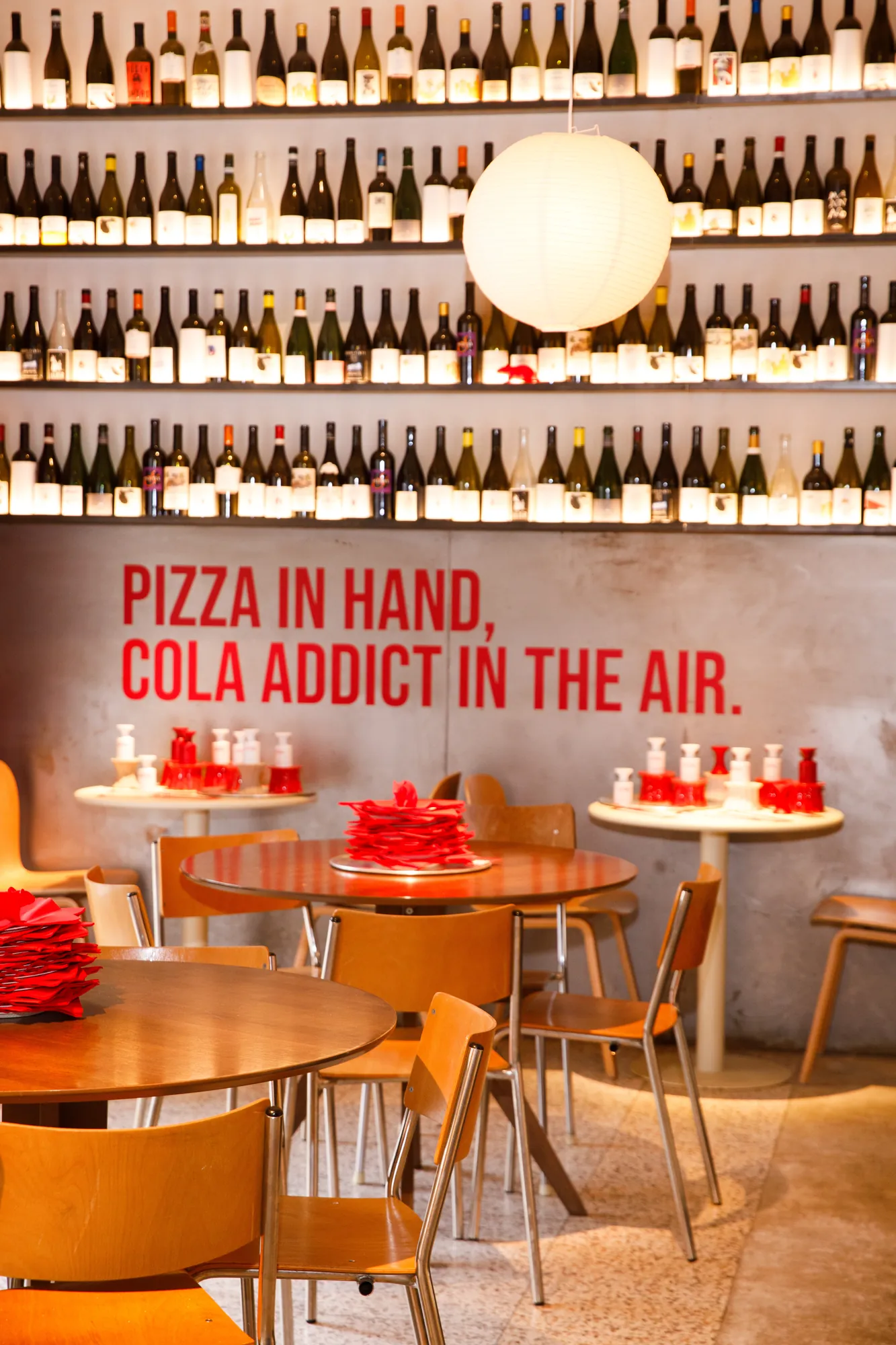 Sala della pizzeria Saya con parete di bottiglie di vino illuminate, tavoli rotondi con tovaglioli rossi e claim a muro "Pizza in hand, cola addict in the air." / Dining room at Saya pizzeria with a lit wall of wine bottles, round tables with red napkins and the wall claim "Pizza in hand, cola addict in the air."