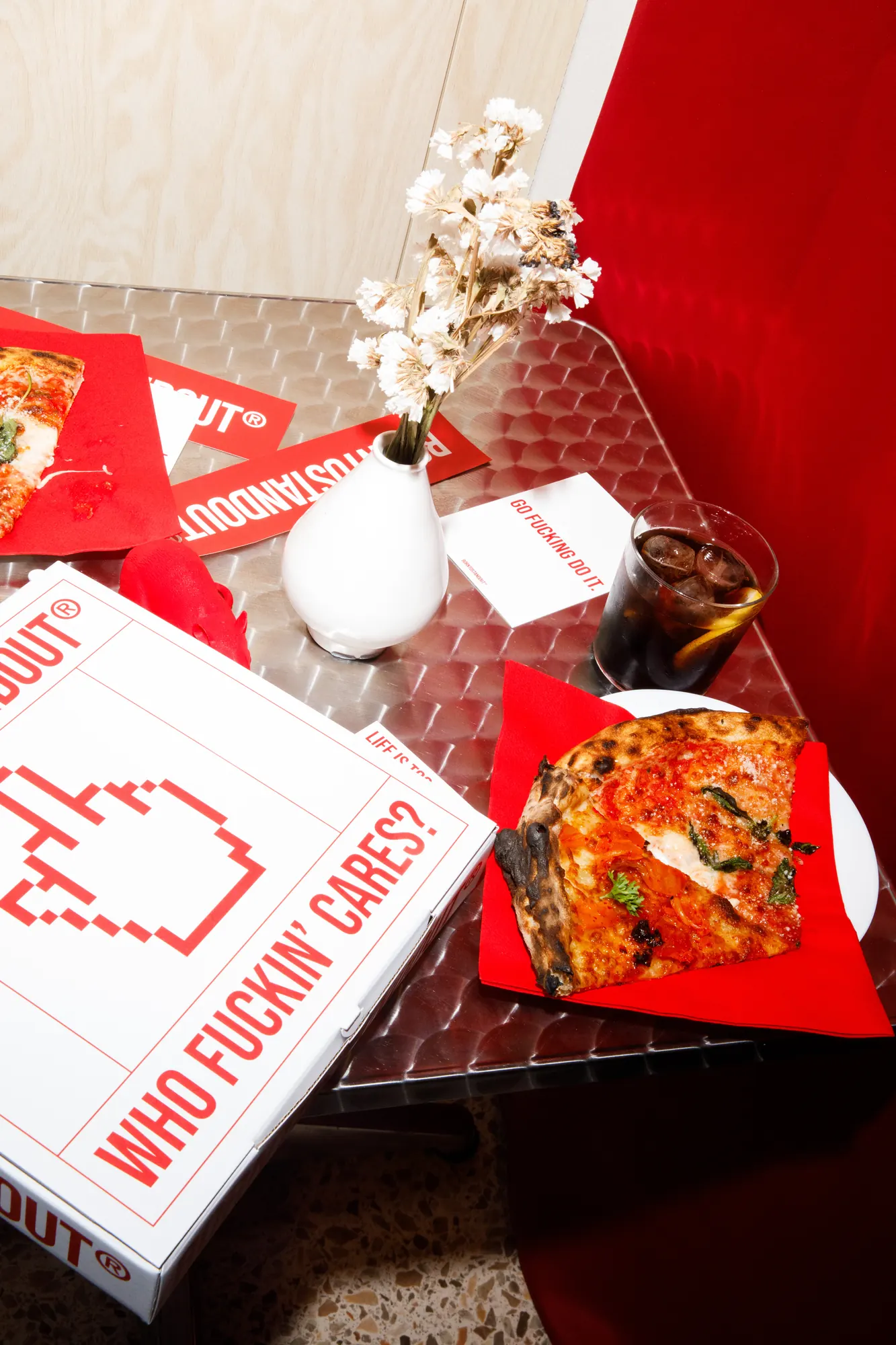 Tavolino in acciaio con trancio di pizza su tovagliolo rosso, bicchiere di cola con ghiaccio e scatola pizza BORNTOSTANDOUT con la scritta "Who fuckin' cares?" / Steel table with a pizza slice on a red napkin, a glass of cola with ice and a BORNTOSTANDOUT pizza box reading "Who fuckin' cares?"