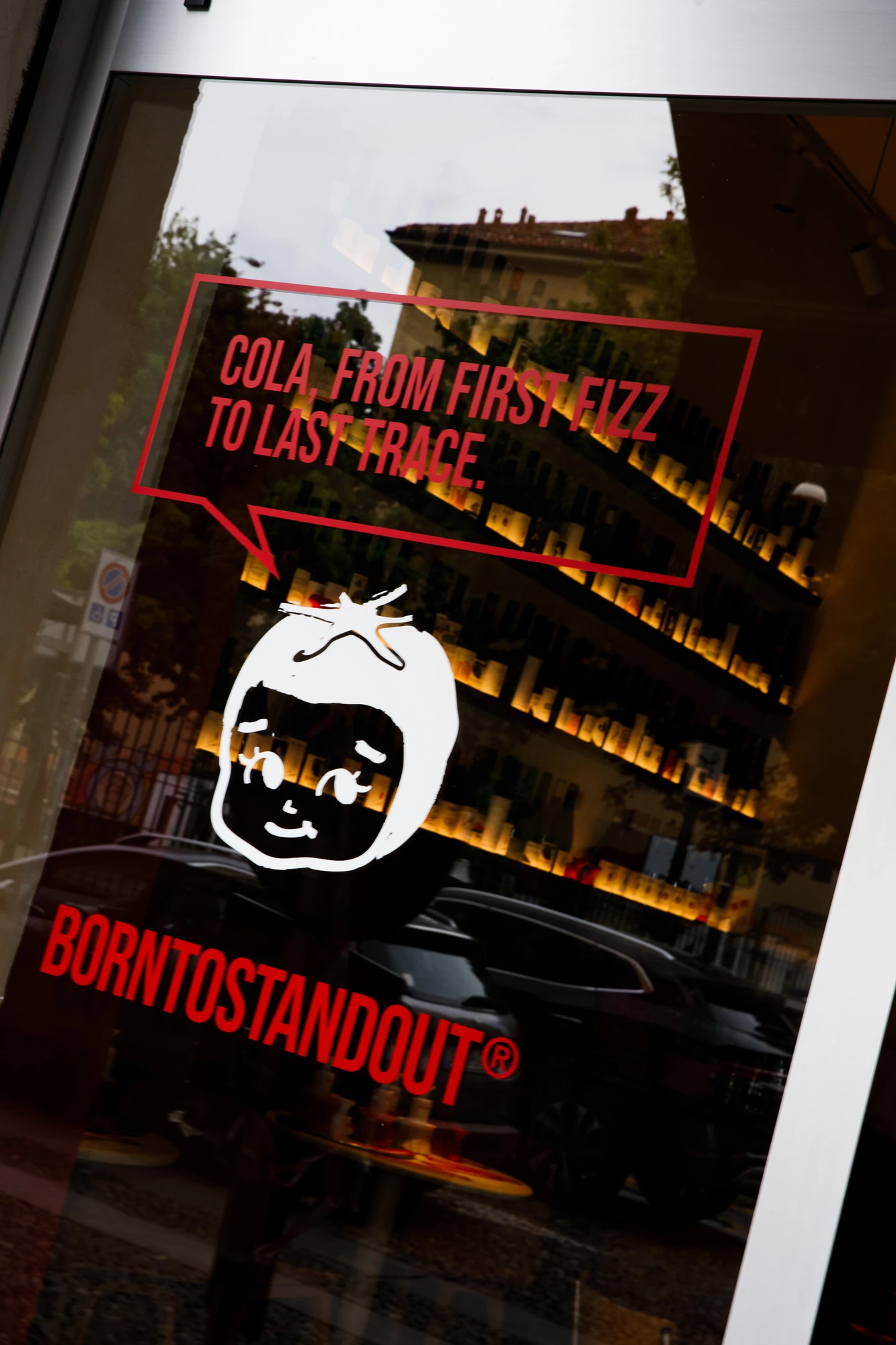 Vetrina della pizzeria con lettering rosso BORNTOSTANDOUT, illustrazione di un pomodoro e il claim "Cola, from first fizz to last trace." / Pizzeria window with red BORNTOSTANDOUT lettering, a tomato illustration and the claim "Cola, from first fizz to last trace."