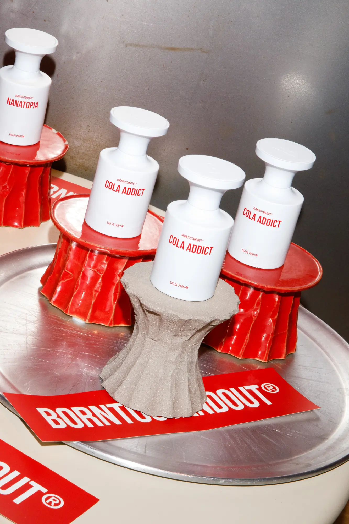 Flaconi di eau de parfum BORNTOSTANDOUT Cola Addict e Nanatopia esposti su alzatine in ceramica rossa sopra un vassoio d'acciaio / BORNTOSTANDOUT Cola Addict and Nanatopia eau de parfum bottles displayed on red ceramic stands on a steel tray