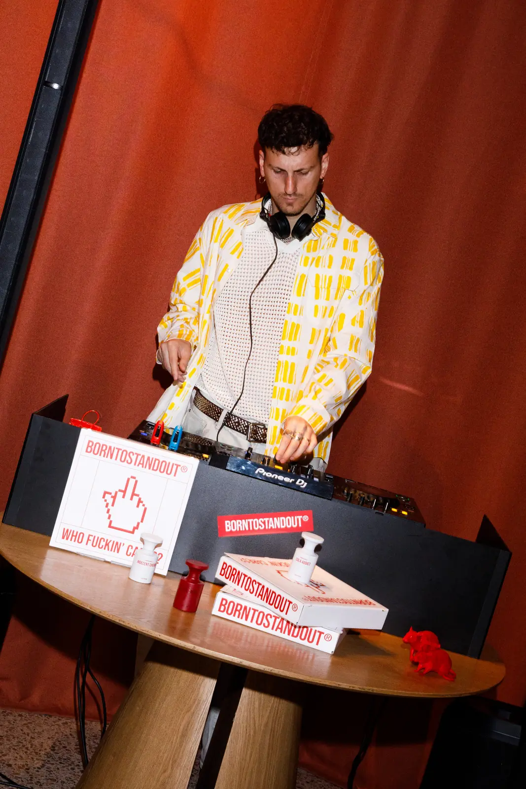 DJ set durante il Craving Club, con consolle appoggiata a un tavolino tra scatole pizza e profumi BORNTOSTANDOUT, davanti a una tenda rossa / DJ set during the Craving Club, console resting on a small table among BORNTOSTANDOUT pizza boxes and fragrances, against a red curtain