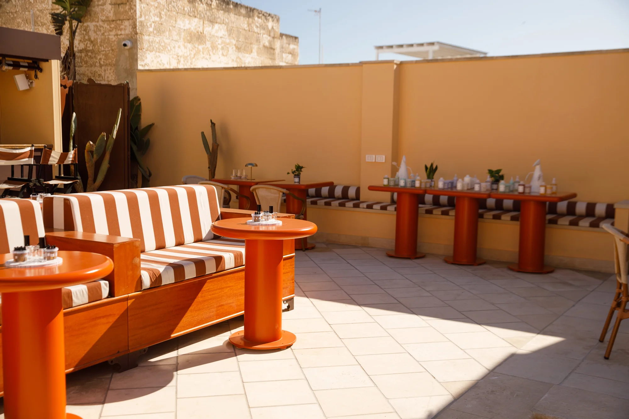 Terrazza esterna dell'evento Davines in Puglia con divani a righe bianche e ruggine, tavolini arancioni e prodotti beauty allineati su una mensola, sotto il sole / Outdoor terrace at the Davines event in Puglia with white and rust striped sofas, orange side tables and beauty products lined up on a shelf, in full sunlight