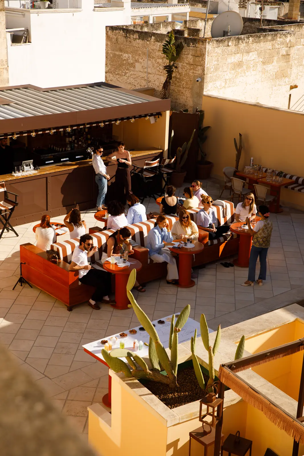 Vista dall'alto della terrazza al tramonto con ospiti seduti sui divani a righe che chiacchierano e bevono, bancone bar sullo sfondo e fichi d'India in primo piano / Aerial view of the rooftop terrace at sunset with guests sitting on striped sofas chatting and drinking, bar counter in the background and prickly pear cactus in the foreground