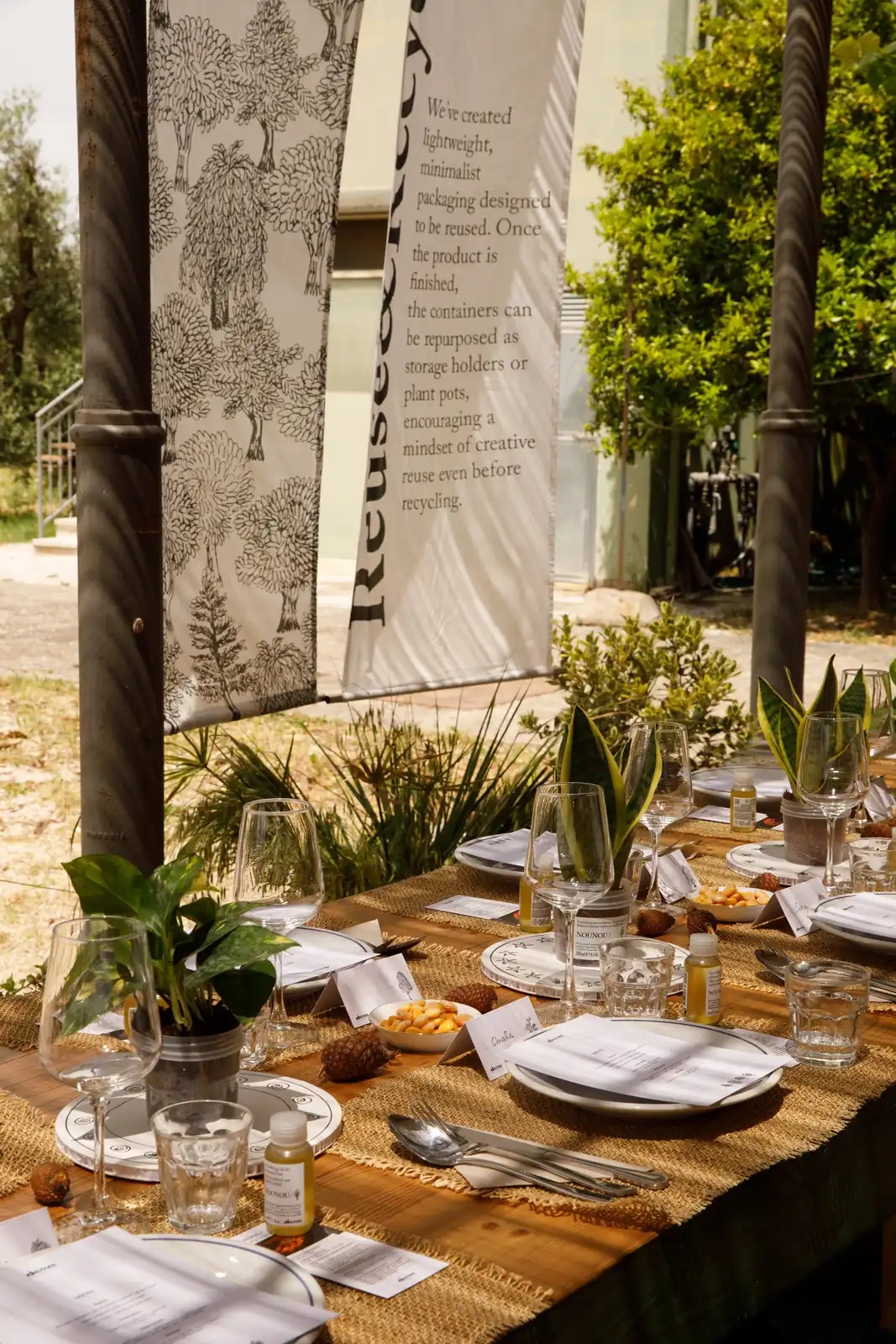 Tavolo conviviale apparecchiato all'aperto tra gli ulivi con runner in juta, piante in vaso e miniature Davines, sotto uno striscione con la scritta "Reuse & Recycle" / Communal table set outdoors among olive trees with jute runners, potted plants and Davines miniatures, beneath a banner reading "Reuse & Recycle"