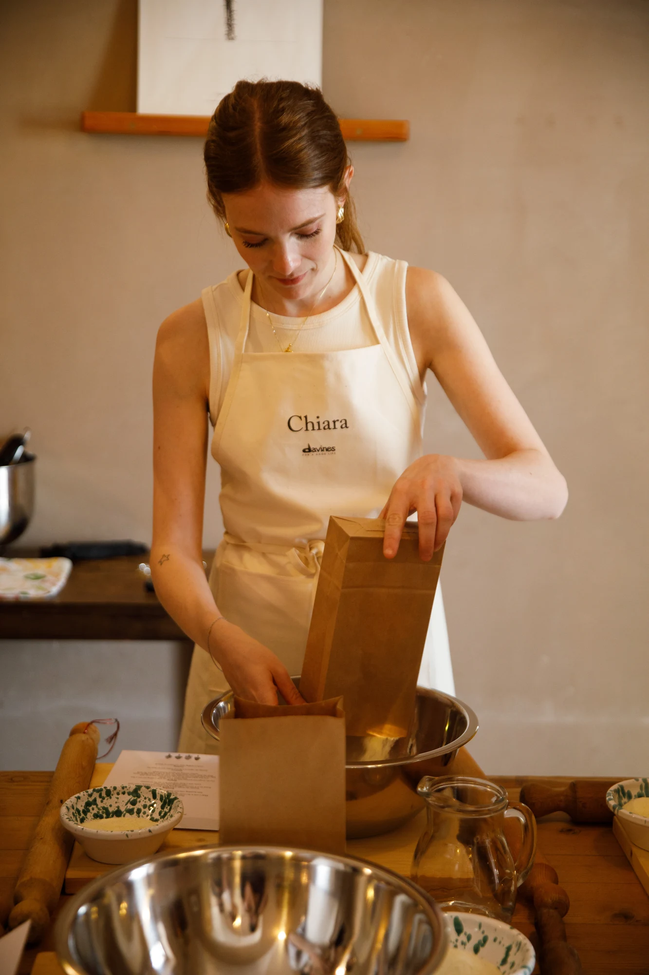 Ragazza con grembiule Davines personalizzato "Chiara" versa farina in un sacchetto di carta durante un laboratorio di cucina / Young woman wearing a personalized Davines apron reading "Chiara" pouring flour into a paper bag during a cooking workshop