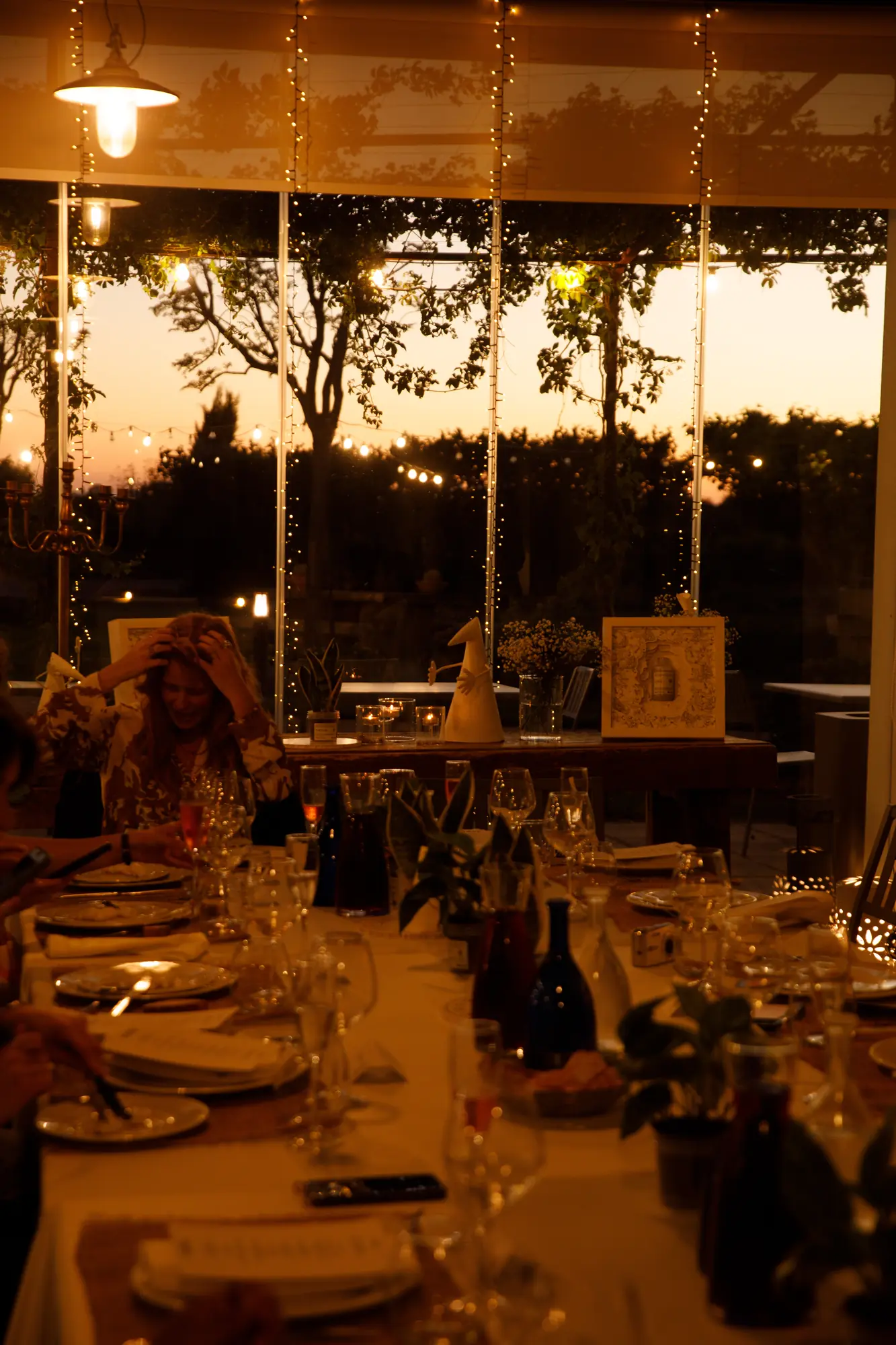 Lunga tavolata apparecchiata al tramonto vista attraverso le vetrate, con luci a filo, candele e ospiti a cena nella campagna pugliese / Long dinner table set at sunset seen through glass walls, with string lights, candles and guests dining in the Apulian countryside