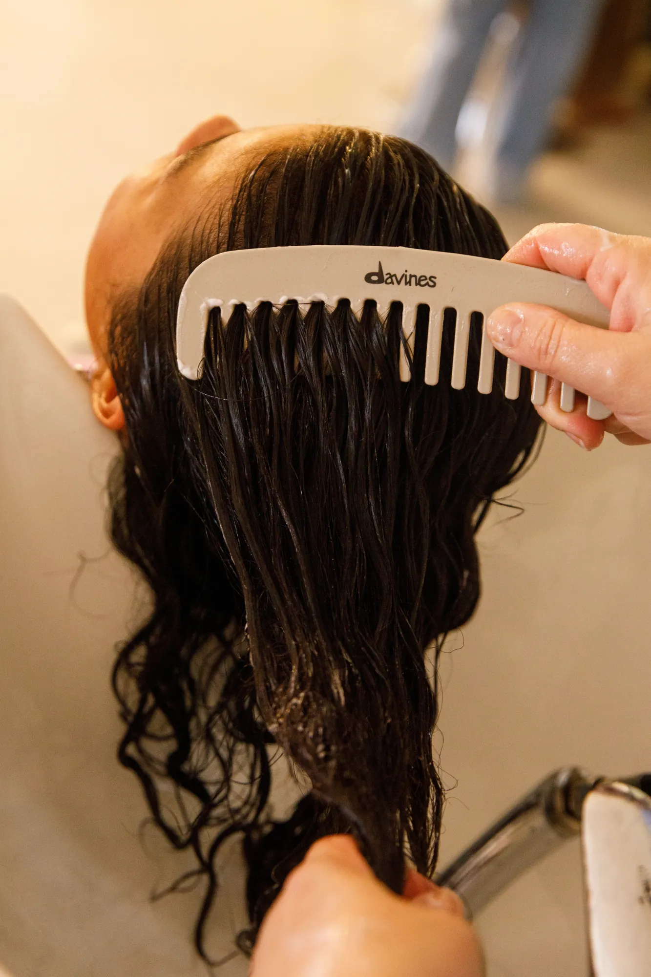 Primo piano di un trattamento capelli: mani che passano un pettine Davines tra i capelli bagnati di una cliente al lavatesta / Close-up of a hair treatment: hands running a Davines comb through a client's wet hair at the salon washbasin