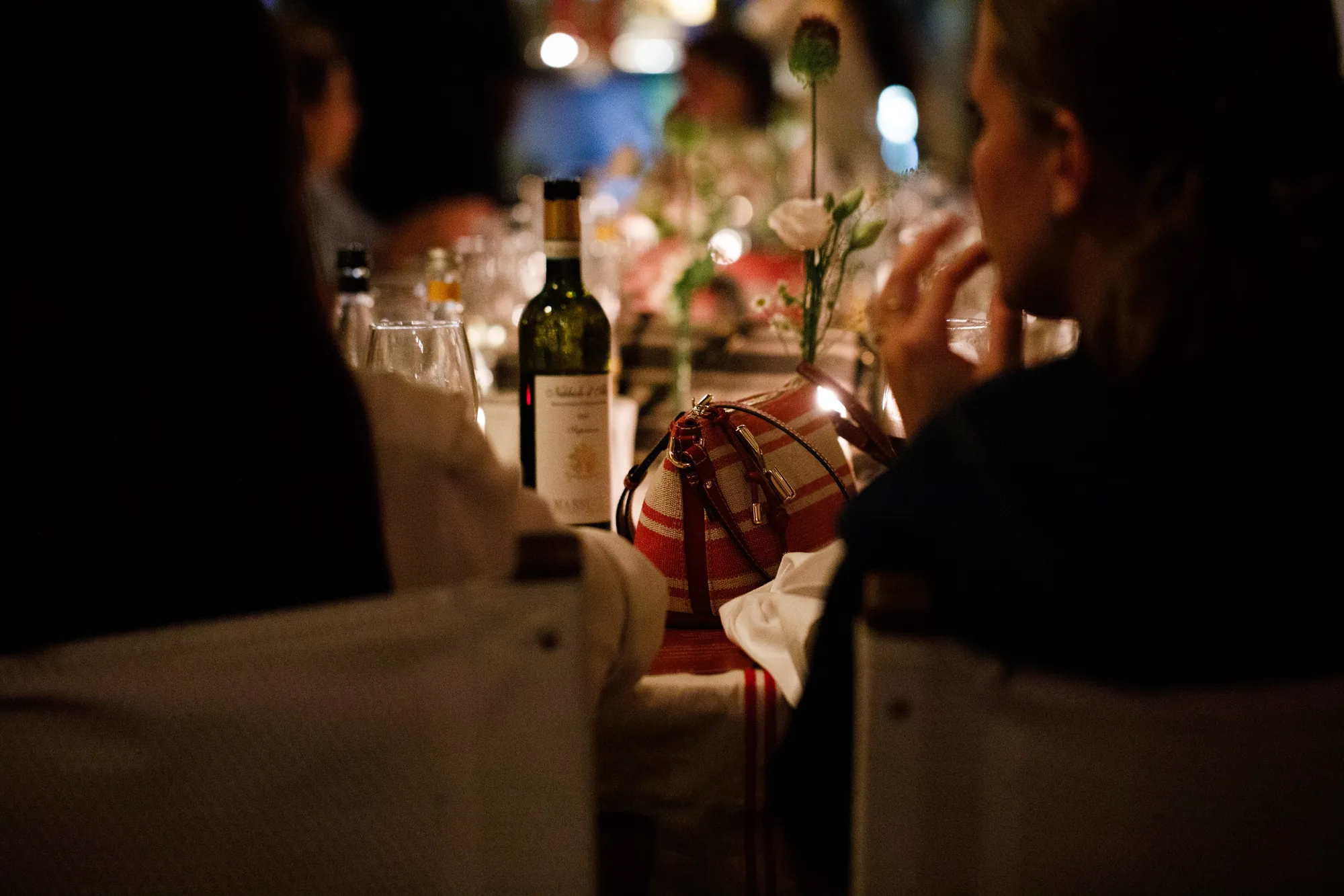 Scorcio della cena Furla Small Talks a lume di candela, con una borsa a righe rosse e bianche appoggiata sulla tavola tra bicchieri e fiori / Candlelit glimpse of the Furla Small Talks dinner, with a red and white striped bag resting on the table among glasses and flowers