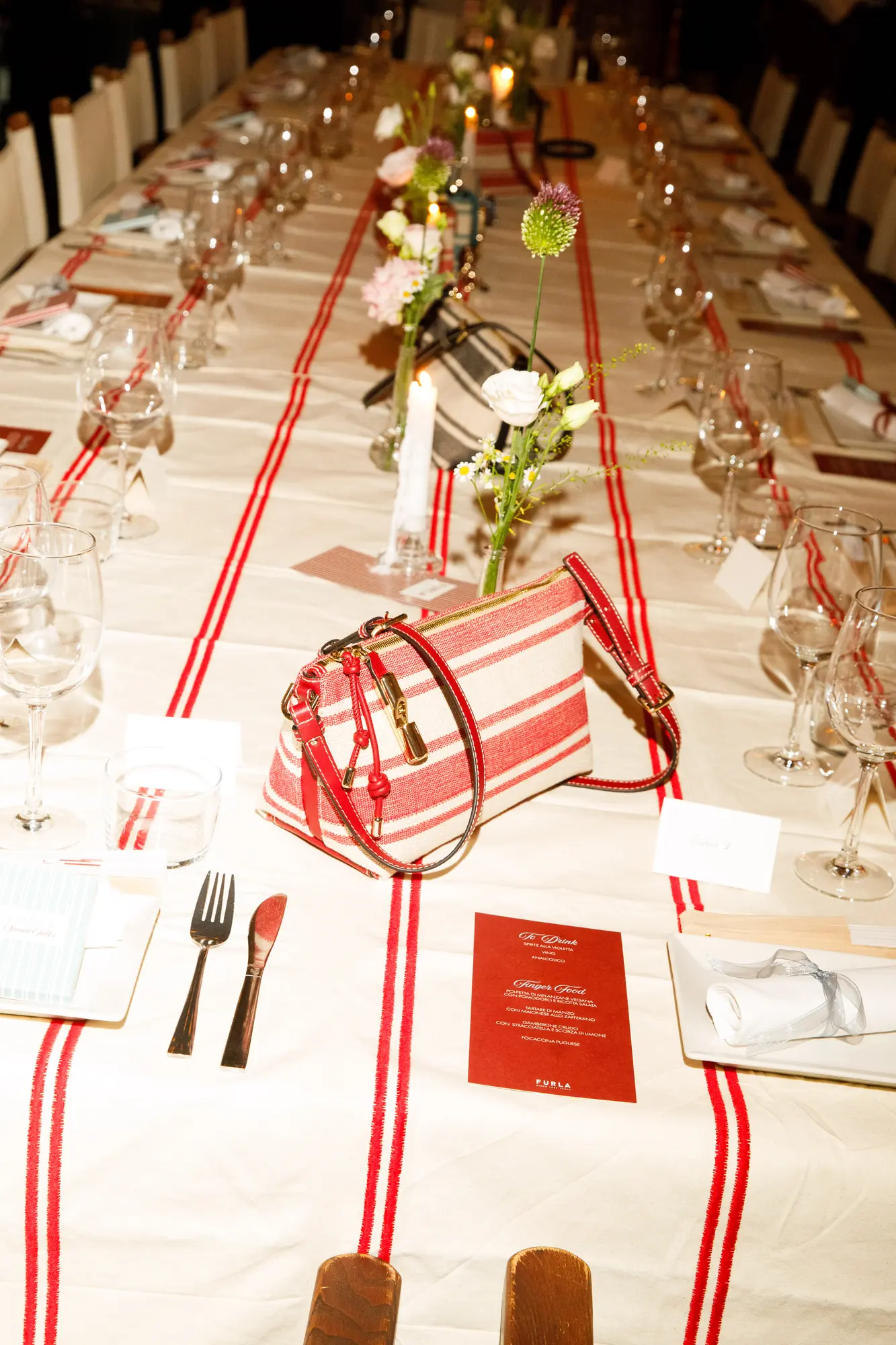 Tavolata imbandita per la cena Furla Small Talks con tovaglia a righe rosse, candele, fiori di campo e una borsa a righe rosse e bianche al centro accanto al menu / Long table set for the Furla Small Talks dinner with red striped linen, candles, wildflowers and a red and white striped bag at the centre next to the menu