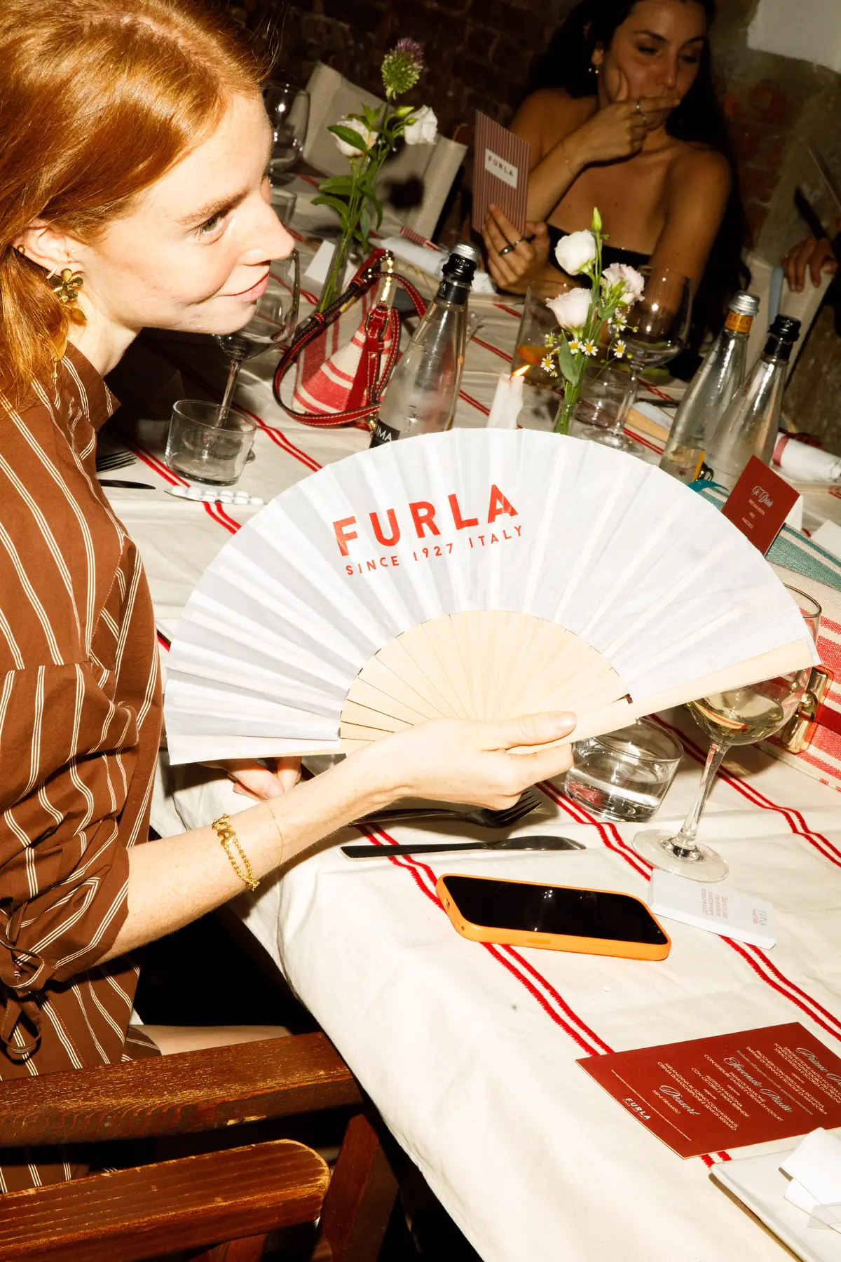 Ospite seduta a tavola sventola un ventaglio bianco con logo "Furla since 1927 Italy", tra bicchieri, menu e una borsa a righe rosse / Guest seated at the table waving a white fan printed with the "Furla since 1927 Italy" logo, among glasses, menus and a red striped bag