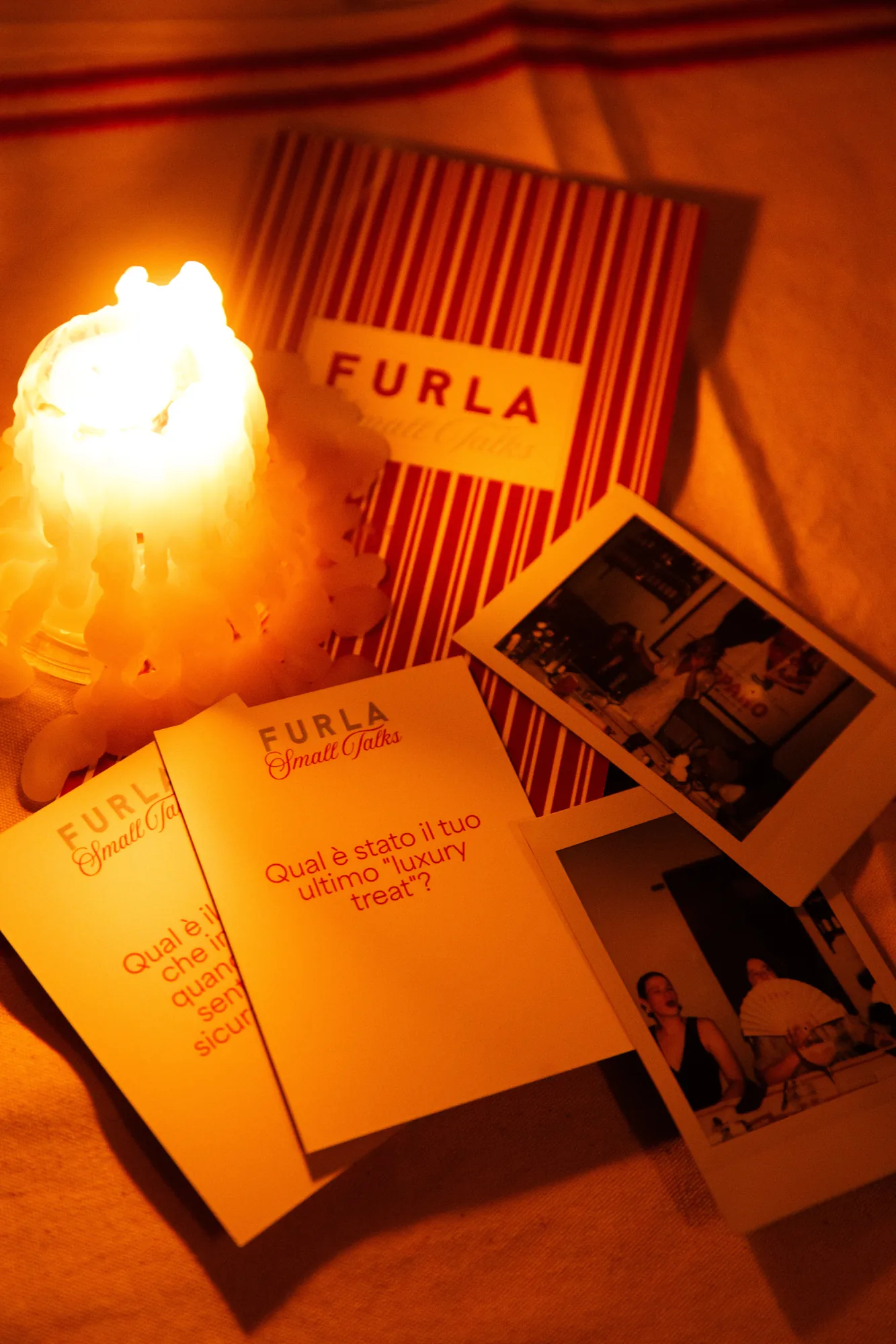 Cartoncini Furla Small Talks con domande stampate, foto istantanee e una candela accesa appoggiati sulla tovaglia a righe rosse / Furla Small Talks conversation cards with printed questions, instant photos and a lit candle resting on the red striped tablecloth