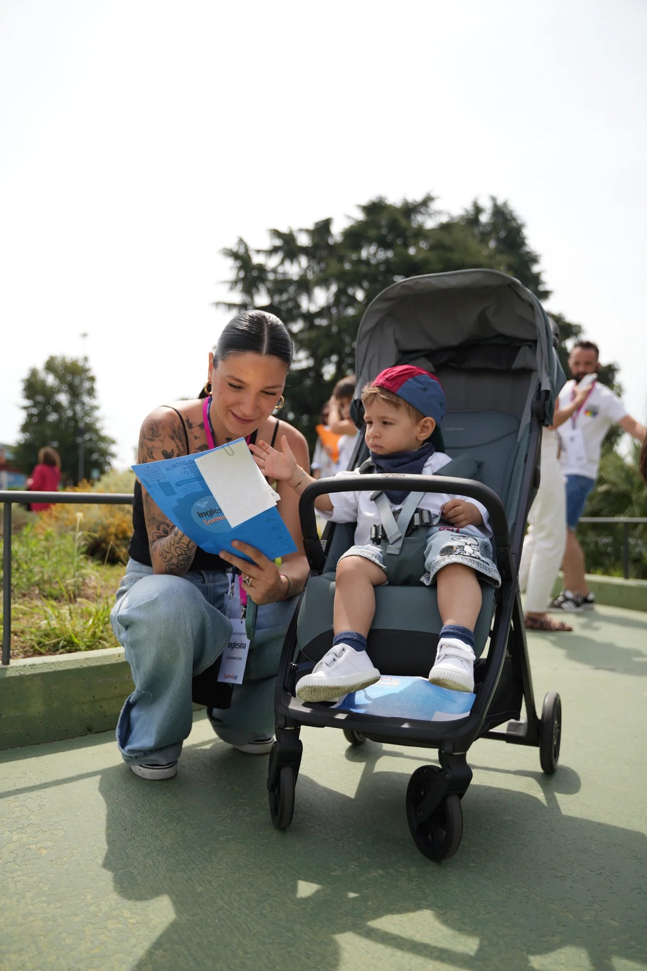 Donna accovacciata accanto a un bambino seduto in un passeggino Inglesina mentre sfoglia una brochure dell'evento, lungo un vialetto del parco Leolandia / Woman crouching next to a toddler seated in an Inglesina stroller while browsing an event brochure, along a walkway at Leolandia park