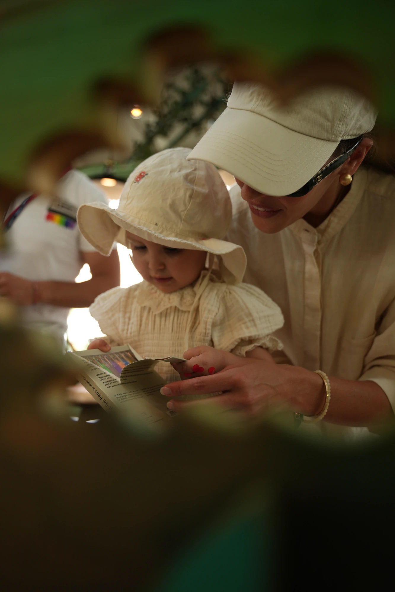 Mamma con visiera bianca e bambina con cappellino di cotone leggono insieme un dépliant durante l'evento Inglesina QUID³ / Mother wearing a white visor and little girl in a cotton sun hat reading a leaflet together during the Inglesina QUID³ event
