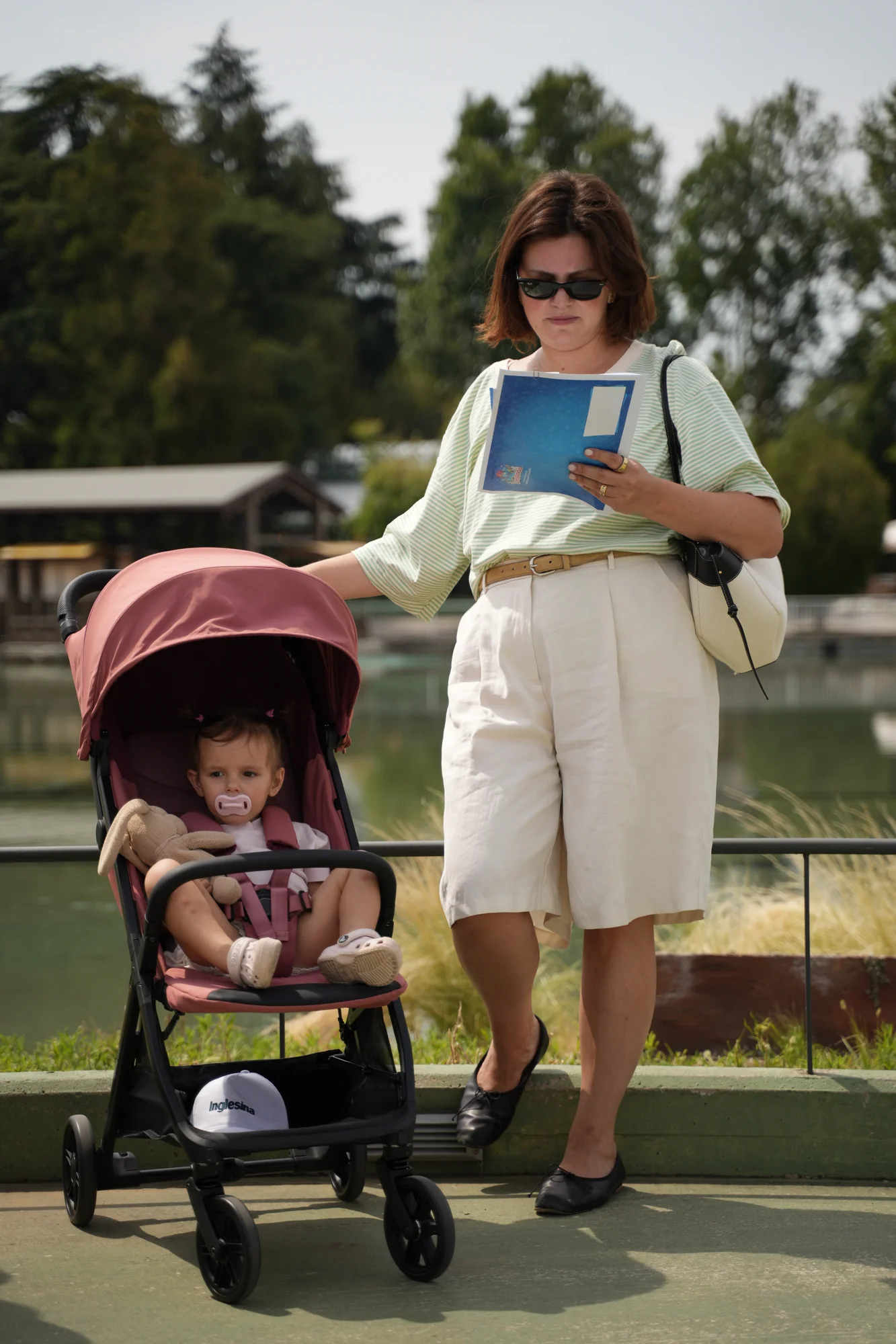 Donna con occhiali da sole spinge un passeggino Inglesina rosa con una bambina a bordo, davanti al laghetto del parco Leolandia / Woman in sunglasses pushing a pink Inglesina stroller with a little girl on board, in front of the lake at Leolandia park