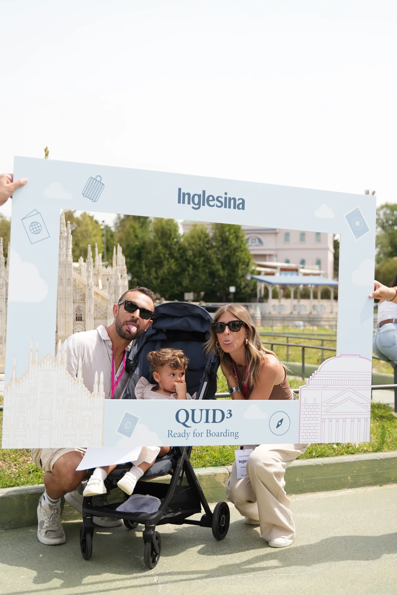 Famiglia con bambino nel passeggino posa dietro la cornice photo booth Inglesina "QUID³ Ready for Boarding", davanti alla miniatura del Duomo di Milano / Family with a child in the stroller posing behind the Inglesina "QUID³ Ready for Boarding" photo booth frame, in front of the miniature Milan Cathedral