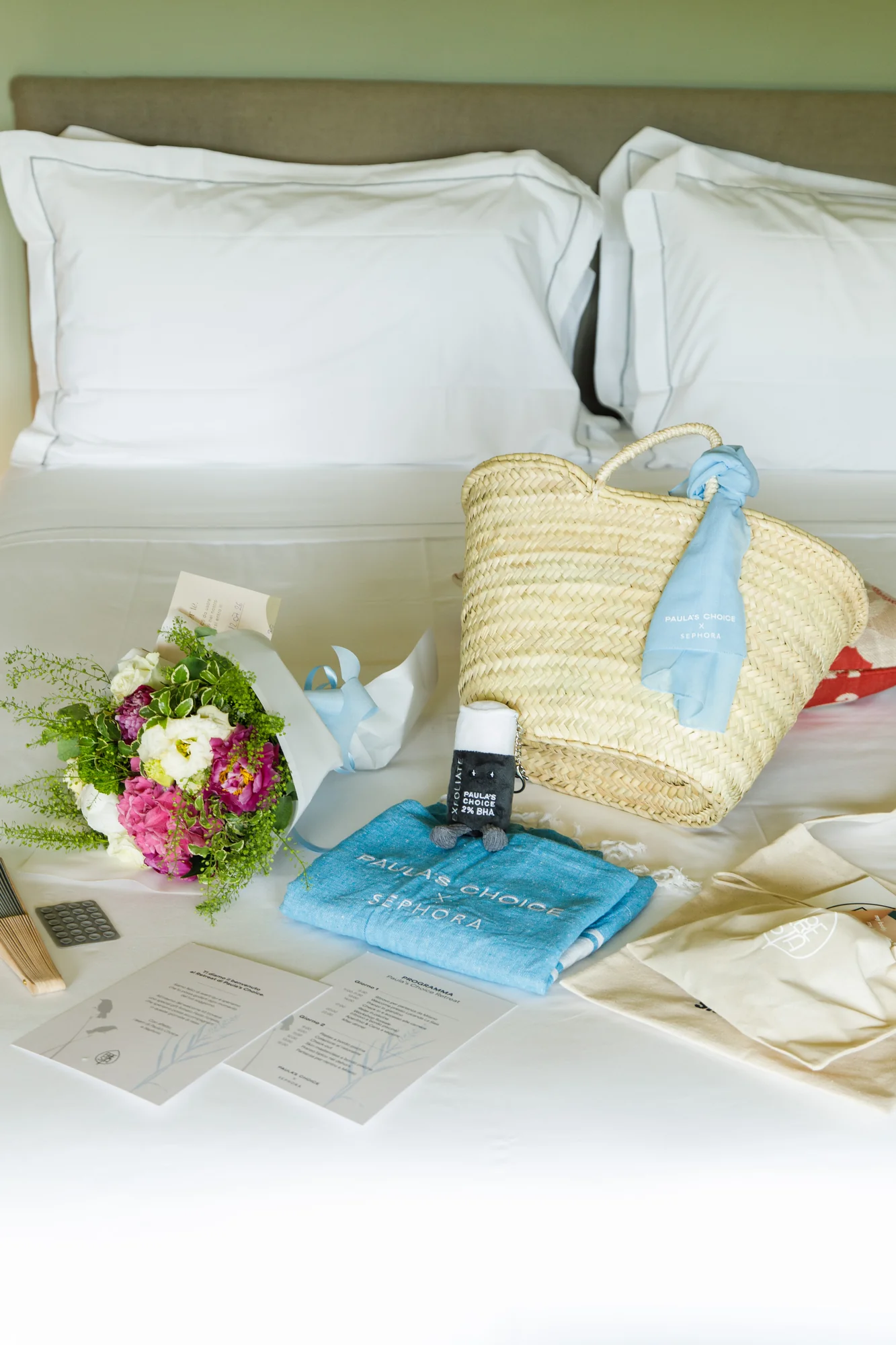 Welcome kit Paula's Choice x Sephora disposto sul letto della camera: borsa in paglia, telo azzurro con logo, bouquet di fiori e programma del retreat / Paula's Choice x Sephora welcome kit laid out on the bed: straw tote, light blue logo towel, flower bouquet and the retreat programme