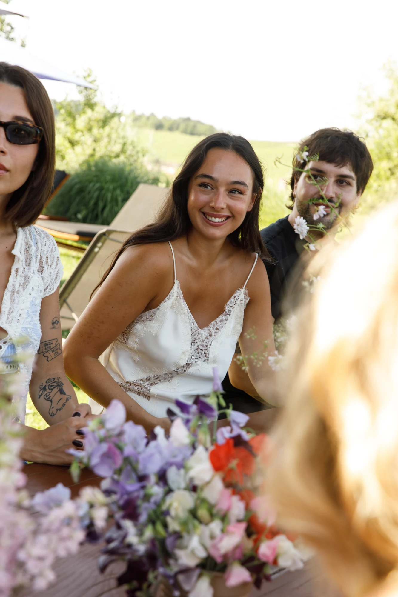 Ospiti sorridenti sedute al tavolo all'aperto tra i vigneti, con un bouquet di fiori colorati sfocato in primo piano / Smiling guests seated at the outdoor table among the vineyards, with a bouquet of colourful flowers blurred in the foreground
