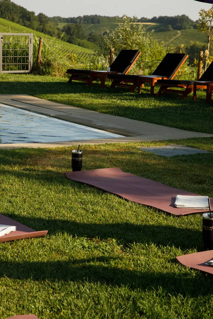 Tappetini da yoga stesi sul prato accanto alla piscina con bicchieri termici Paula's Choice, davanti alle colline e ai vigneti / Yoga mats laid out on the lawn beside the pool with Paula's Choice tumblers, facing the hills and vineyards