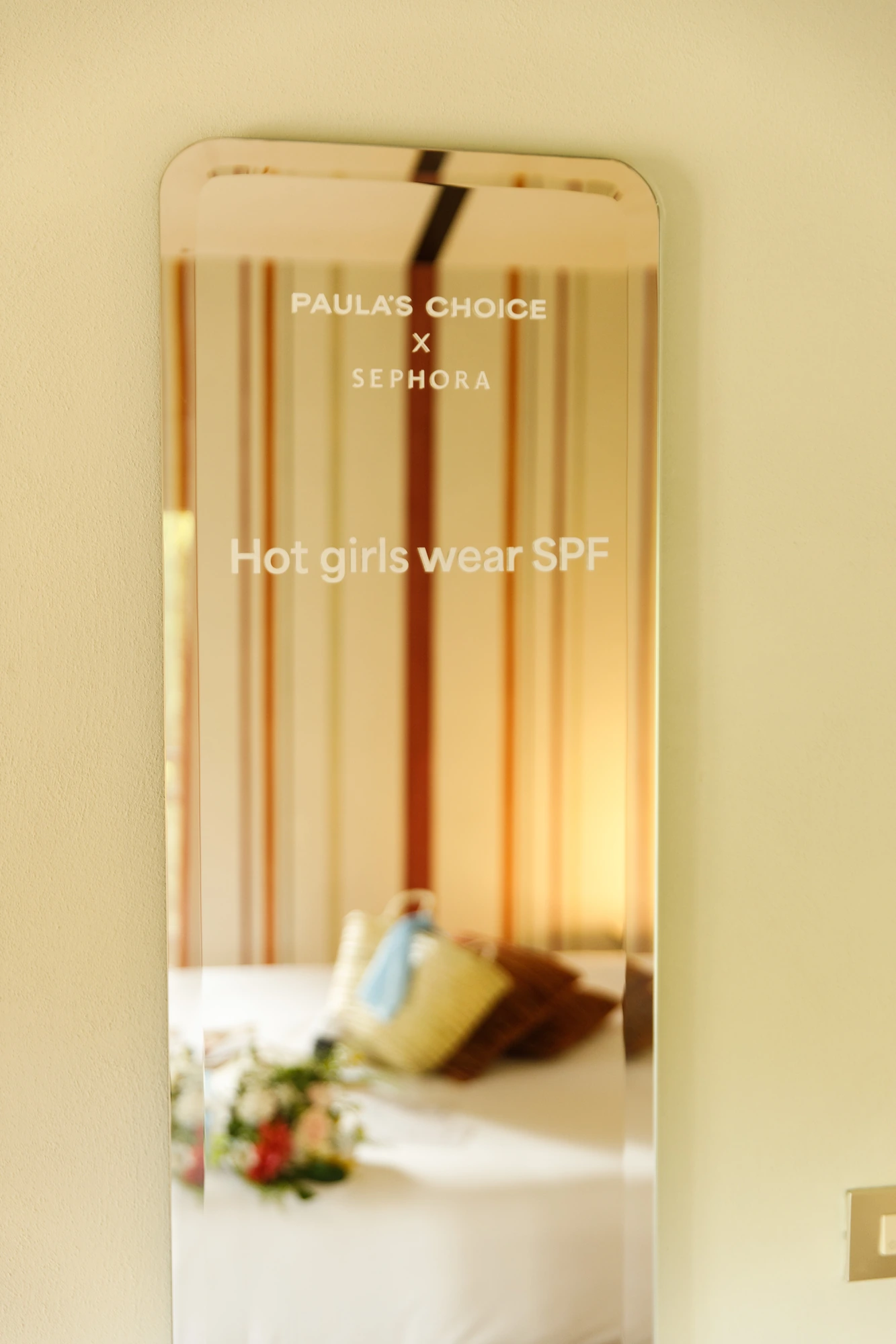 Specchio a parete con incisione "Paula's Choice x Sephora – Hot girls wear SPF" che riflette il welcome kit sul letto / Wall mirror engraved "Paula's Choice x Sephora – Hot girls wear SPF" reflecting the welcome kit on the bed