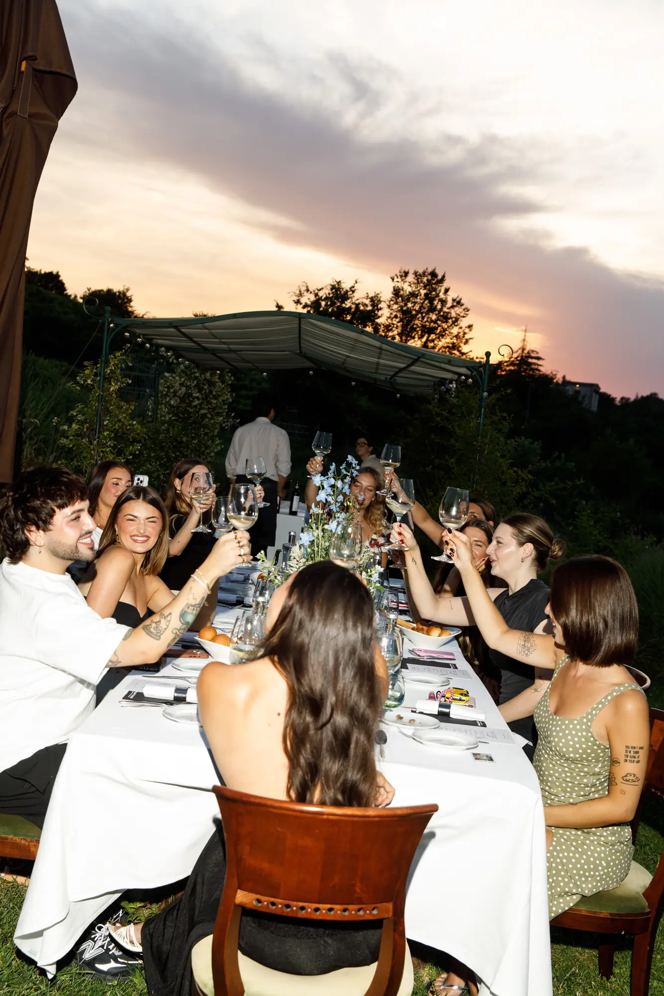 Brindisi degli ospiti alla cena all'aperto del retreat, con calici alzati sopra la tavola bianca e cielo al tramonto sullo sfondo / Guests toasting at the retreat's outdoor dinner, glasses raised above the white table with a sunset sky in the background