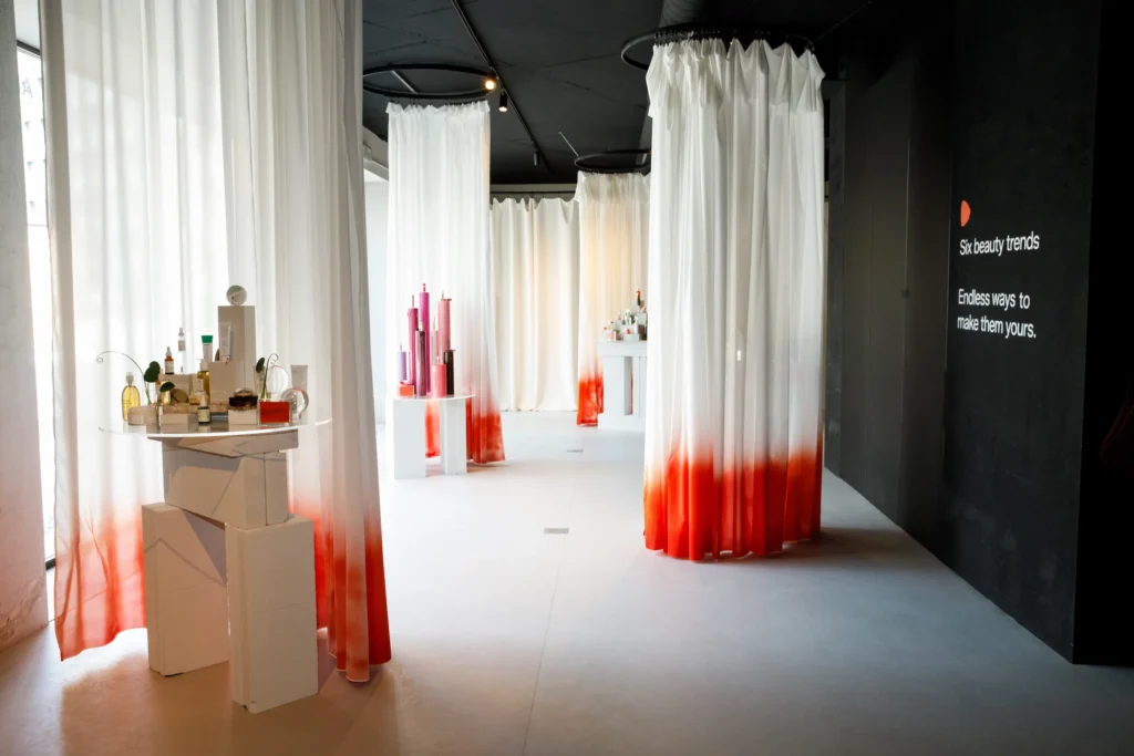 Allestimento espositivo beauty con tende bianche sfumate di rosso appese a strutture circolari e prodotti cosmetici su piedistalli bianchi, accanto a una parete nera con la scritta "Six beauty trends" / Beauty exhibition setup with white curtains dip-dyed in red hanging from circular frames and cosmetic products on white pedestals, next to a black wall reading "Six beauty trends"